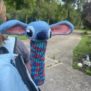 May include: A blue and pink crochet Stitch doll with large ears and a cylindrical body. The doll is held by a person wearing a light blue backpack. The background features a paved path, trees, and greenery.