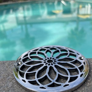 May include: A silver, round, decorative drain cover with a geometric, floral design. The drain cover is sitting on a gray, textured surface.