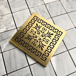 May include: A square, brushed gold-tone metal drain cover featuring a central cutout design of symmetrical, leaf-like patterns surrounded by a continuous geometric Greek key border. The drain cover is positioned flat on a white marble tile surface.