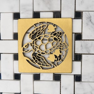 May include: A square, brushed gold-tone metal drain cover featuring a circular cutout design. The intricate pattern depicts a sea turtle swimming alongside two smaller fish, all rendered in a detailed, openwork style within the circular frame.