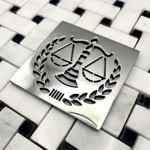 May include: A square, polished silver-tone metal shower drain cover featuring a laser-cut design of a scale of justice centered within a circular laurel wreath. The metallic surface is reflective and smooth, showcasing precise cutouts for drainage.