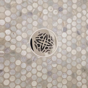 May include: A close-up of a silver drain cover with a decorative design, set in a shower floor with small, round, gray and white tiles.