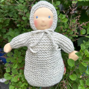 Puede incluir: Muñeca hecha a mano con un conjunto y capucha de punto gris claro. La muñeca tiene una cara color melocotón con ojos bordados azules y mejillas rosadas. Los brazos y las manos de la muñeca son de color beige claro. La muñeca está sobre un fondo de follaje verde.