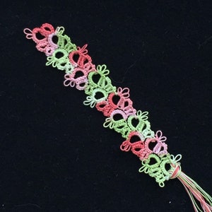 May include: A handmade, colorful tatting lace bookmark. The bookmark features a repeating floral design in shades of pink, green, and light pink. The bookmark has a tassel of matching threads at the end. The background is black.