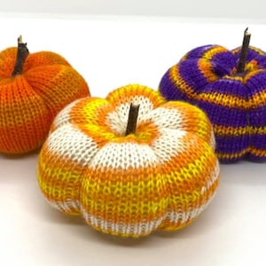 May include: Three knitted pumpkins in orange, white, and purple with yellow stripes. The pumpkins are handmade and have a rustic, autumnal feel.