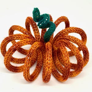 May include: A handmade, knitted pumpkin in shades of orange and brown, with a green stem. The pumpkin is made of looped yarn, creating a three-dimensional effect. The stem is a tightly wound, dark green yarn.