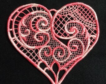 May include: A pink and white heart-shaped lace applique with intricate swirls and a delicate design.