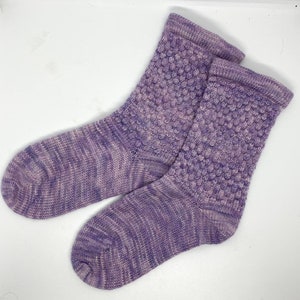 May include: A pair of hand-knitted socks in shades of purple and lavender. The socks feature a textured, bubble-like pattern on the upper portion and a ribbed cuff. The yarn has a variegated color effect, creating a unique look.