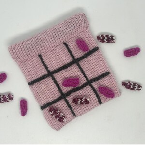 May include: A pink knitted bag with a black tic-tac-toe board design. The bag is surrounded by small knitted pieces in various shades of pink and purple.