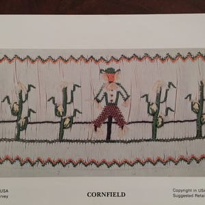 May include: A white fabric embroidery pattern with a scarecrow in a cornfield. The scarecrow is wearing a brown hat, a blue shirt, red pants, and a brown belt. The corn stalks are green with yellow ears of corn. The pattern is surrounded by a red and green border.