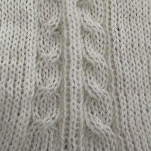 May include: Close-up of a white knitted fabric with a cable knit design. The pattern features vertical columns of knit stitches, with a central cable design. The texture is soft and the color is a bright white.