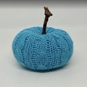 May include: A round, light blue knitted pumpkin decoration featuring a textured cable knit pattern. The top of the pumpkin is adorned with a short, brown, natural wooden twig that serves as a stem, contrasting with the soft blue yarn.