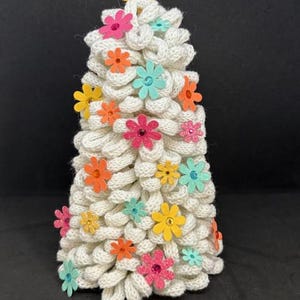 May include: A decorative, handmade, white knitted tree adorned with colorful, glittery flower embellishments. The flowers are in shades of pink, orange, yellow, and teal. The tree is set against a black background.