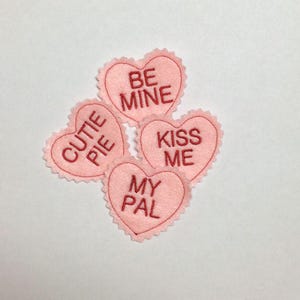 May include: Four light pink heart-shaped felt patches with scalloped edges. Each patch features red embroidered text: BE MINE, CUTIE PIE, KISS ME, and MY PAL. The patches are arranged in a cluster, showcasing the soft texture and stitching.