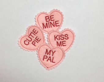 Könnte beinhalten: Vier hellrosa herzförmige Filzaufnäher mit gezackten Rändern. Jeder Aufnäher trägt einen roten Sticktext: BE MINE, CUTIE PIE, KISS ME und MY PAL. Die Aufnäher sind in einer Gruppe angeordnet und zeigen die weiche Textur und die Stickerei.