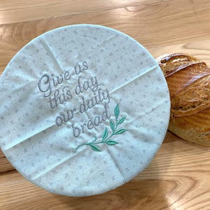 May include: A round, light blue fabric bowl cover with a small, repeating floral pattern. The cover features the embroidered text "Give us this day our daily bread" in dark gray, accompanied by a small, green embroidered leafy branch design.