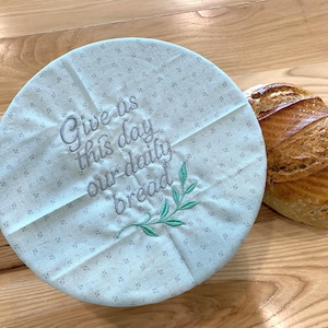 May include: A light blue bread cover with the embroidered text "Give us this day our daily bread" and a green leaf design. The cover is round and has a small floral pattern. A loaf of bread is partially visible.