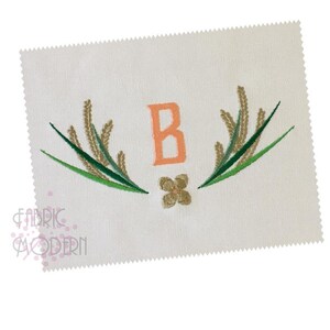 以下が含まれることがあります： ピーチ色の「B」の文字を刺繍したデザインで、緑と金のスタイリッシュな葉と小さな花で囲まれています。