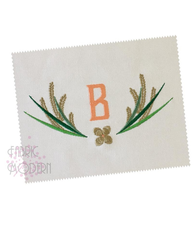 以下が含まれることがあります： ピーチ色の「B」の文字を刺繍したデザインで、緑と金のスタイリッシュな葉と小さな花で囲まれています。