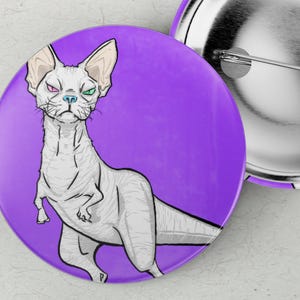 Könnte beinhalten: Ein violetter Button mit einer silbernen Nadel und einer Cartoon-Illustration einer haarlose Katze mit Dinosaurier-Merkmalen. Die Katze hat einen strengen Ausdruck und mehrfarbige Augen. Ein zweiter Button ist sichtbar.