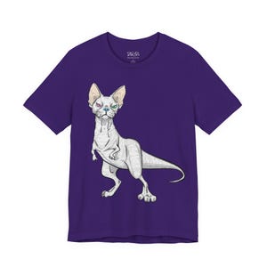 May include: A purple short-sleeved t-shirt featuring a graphic illustration of a hairless cat with a dinosaur-like body, a long tail, and heterochromia with one purple eye and one green eye, centered on the front of the garment.