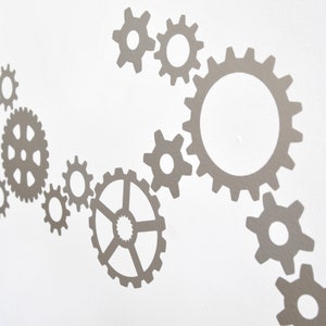 May include: A set of silver metal gears of various sizes arranged in a flowing pattern on a white wall.