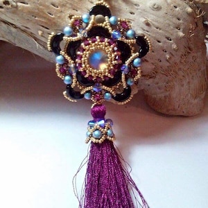 May include: A beaded pendant with a purple tassel. The pendant is made of black, gold, and blue beads and features a iridescent center stone.