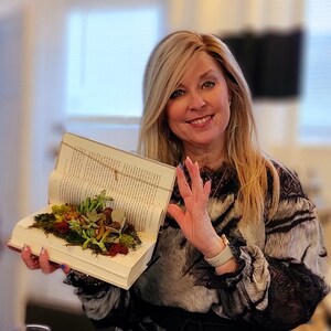 May include: A woman holds open a book with a small succulent garden inside. The book is open to a page with text and the garden is made up of small green plants and moss.