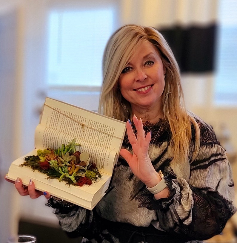 May include: A woman holds open a book with a small succulent garden inside. The book is open to a page with text and the garden is made up of small green plants and moss.