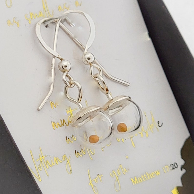 May include: A pair of silver dangle earrings featuring small, clear glass globes. Each globe contains a single, tiny mustard seed. The earrings are attached to a display card with gold script text that reads, "Nothing will be impossible for you. Matthew 17:20."