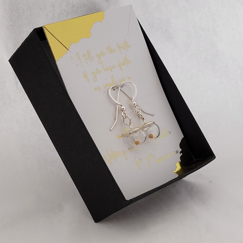 May include: A pair of silver hook earrings featuring small clear glass spheres containing a single mustard seed. The earrings are displayed on a white card with gold foil text that reads, I tell you the truth, if you have faith as small as a mustard seed, you can say to this mountain, move from here to there and it will move. Nothing will be impossible for you. Matthew 17:20, inside a black box.