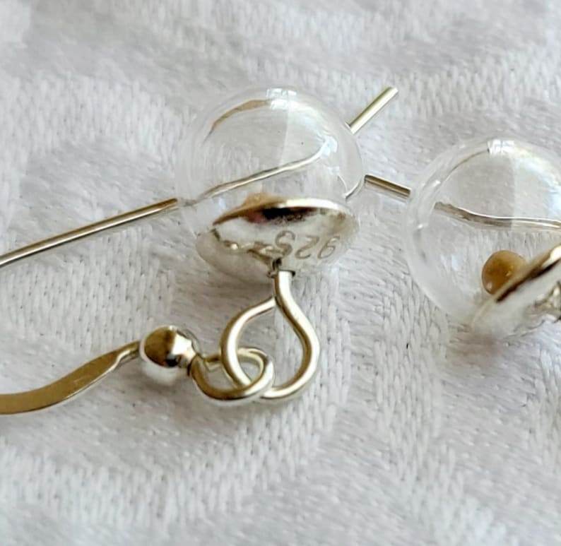 May include: A pair of dangle earrings featuring clear glass spheres attached to silver-toned metal caps. The metal caps are stamped with the text 925, indicating sterling silver composition. Each earring includes a curved silver-toned ear wire hook.