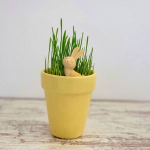 May include: A yellow ceramic pot with green grass growing inside. A small, light brown bunny figurine is sitting in the grass.