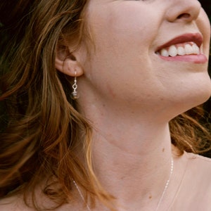 May include: A silver-toned earring featuring a small, clear, spherical glass bead pendant suspended from a simple metal hook. The earring hangs from the earlobe, showcasing a minimalist design with a transparent orb that catches the light.