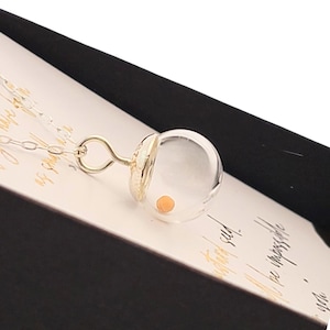 May include: A silver chain necklace featuring a clear, spherical glass pendant. Inside the hollow glass orb sits a single, small, round, light orange bead. The pendant is attached to the chain by a silver loop and a textured silver cap.