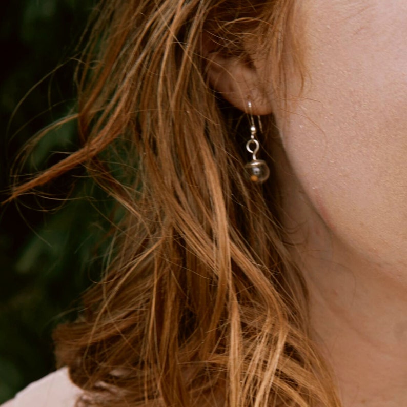 May include: A silver dangle earring featuring a small, clear, spherical glass bead suspended from a thin metal hook. The transparent orb contains a tiny, dark, circular element visible within its center, hanging from a simple silver wire loop.