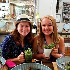 May include: Two women are making their own succulent gardens. One woman is wearing a white hat and a blue floral shirt. The other woman is wearing a pink shirt and a blue lanyard with the word "UNITE" on it. Both women are holding white ceramic cups with succulents planted in them.