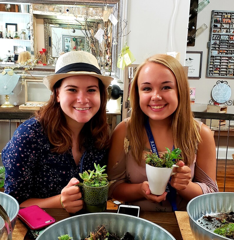 May include: Two women are making their own succulent gardens. One woman is wearing a white hat and a blue floral shirt. The other woman is wearing a pink shirt and a blue lanyard with the word "UNITE" on it. Both women are holding white ceramic cups with succulents planted in them.