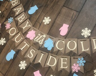 May include: A decorative banner featuring brown cardstock pennants with white lettering that spells out "BABY IT'S COLD OUTSIDE." The banner is accented with light blue and pink paper mittens, white snowflakes, and a pink winter hat cutout.