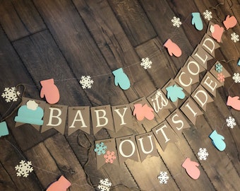 May include: A decorative banner featuring brown pennants with white letters spelling out BABY IT'S COLD OUTSIDE, accented with light blue and pink mitten cutouts, white snowflake shapes, and a light blue winter hat, all strung on twine.