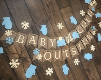 May include: A decorative banner featuring brown cardstock pennants with white lettering that spells out "BABY IT'S COLD OUTSIDE." The banner is accented with light blue paper mittens, white snowflake cutouts, and a light blue winter hat.