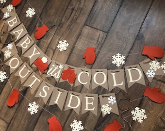 May include: A decorative banner featuring brown cardstock pennants with white lettering that spells out "BABY IT'S COLD OUTSIDE." The banner is accented with red textured mitten cutouts and white snowflake shapes strung along a thin twine cord.