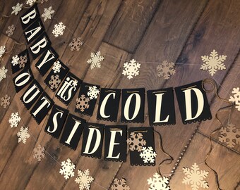 May include: A decorative banner featuring black rectangular cardstock flags with cream-colored lettering that spells out "BABY IT'S COLD OUTSIDE." The banner is strung on twine and accented with numerous die-cut cream and tan paper snowflakes.