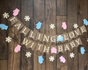 May include: A decorative banner featuring brown pennants with white lettering that spells out FALLING IN LOVE WITH BABY. The banner is accented with light pink and light blue mitten cutouts, white snowflake shapes, and a small blue hat icon.