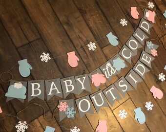May include: A decorative banner featuring grey pennants with white lettering that spells out BABY ITS COLD OUTSIDE, accented with light blue and pink paper cutouts of mittens and white snowflakes strung together on a thin brown twine cord.