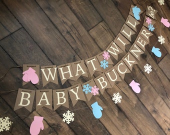 May include: A two-tiered banner made of brown cardstock pennants featuring the white text "WHAT WILL BABY BUCKNER BE?" accented with light blue and pink paper mittens and white snowflake cutouts, all strung together on a thin brown twine cord.