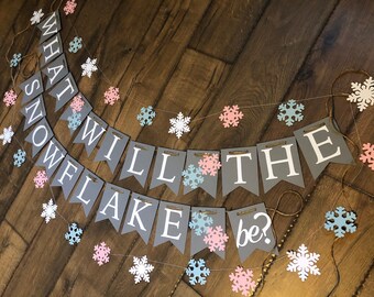 May include: A decorative banner featuring grey pennants with white lettering that spells out "WHAT WILL THE SNOWFLAKE be?" strung on twine. The banner is accented with numerous small, die-cut snowflakes in shades of white, light blue, and soft pink.