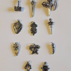 May include: A collection of 10 silver charms featuring various fantasy and mythical designs, including a sword, a hammer, a lion, an eye, a shield, a skull, a horn, and two symbols.