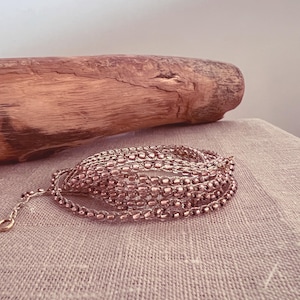 Può includere: Una collana a catena con perline color oro rosa composta da più fili di piccole perline metalliche uniformi disposte in una formazione circolare avvolta, completa di una delicata catenella di estensione e una piccola chiusura circolare.