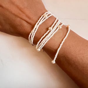 May include: White beaded wrap bracelet with multiple strands, accented with small pearls. The bracelet is wrapped around a wrist, showcasing its delicate design and texture. A simple and elegant accessory.
